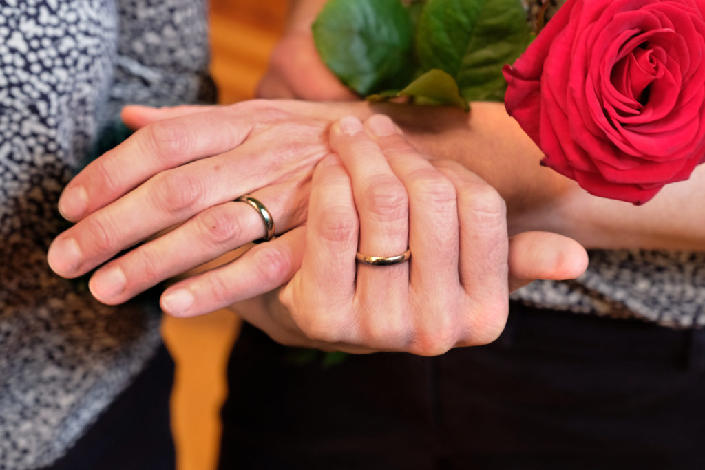 Zwei sich halttende Hände mit Eheringen und einer roten Rose